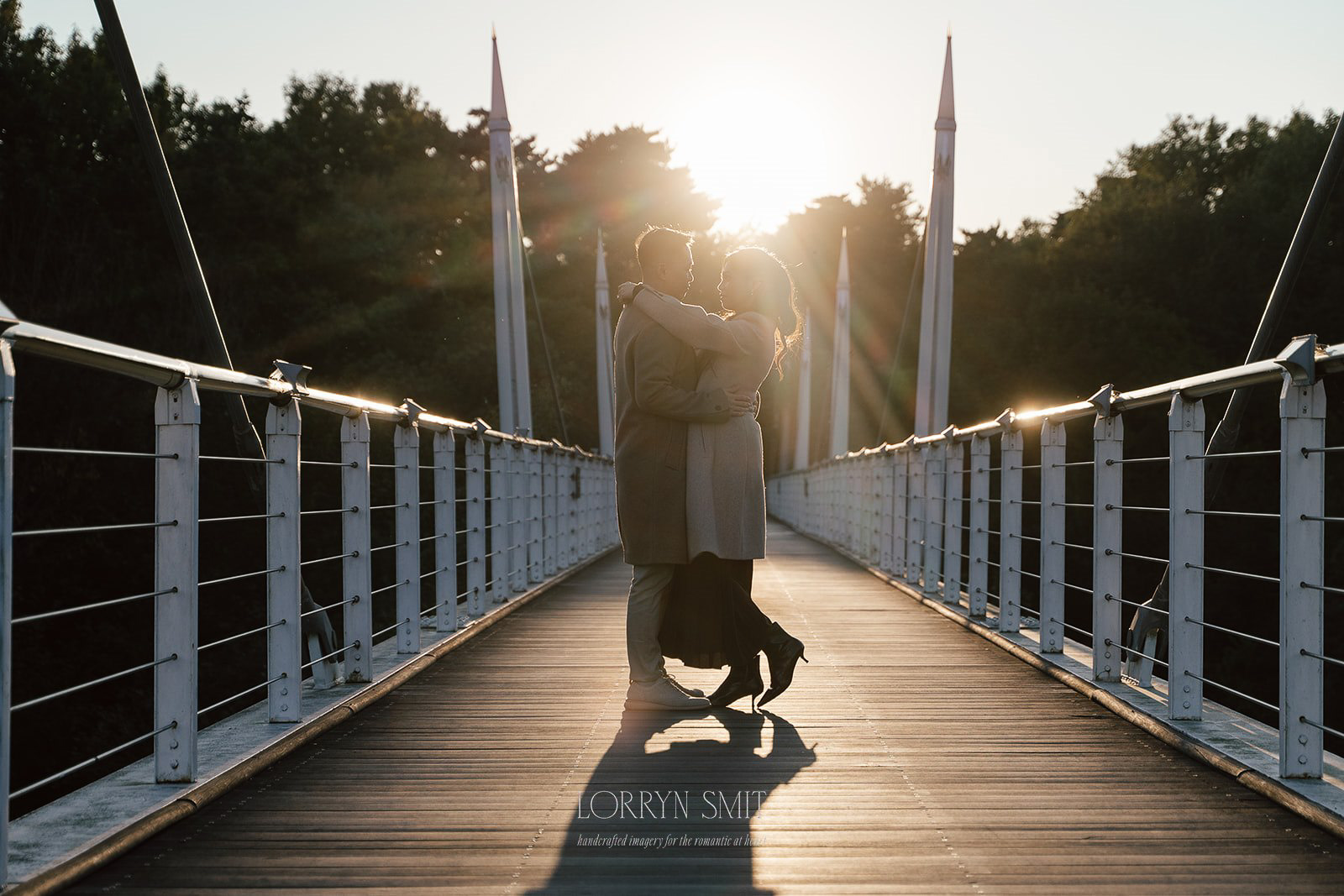 A couple stands embracing on a pedestrian bridge at sunset, silhouetted against the sun with trees in the background.