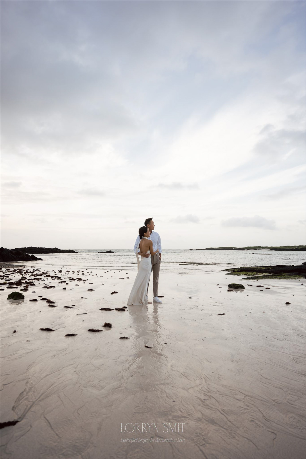 A couple stands together on a wet sandy beach, facing the ocean under a cloudy sky, with rocks and seaweed scattered around during their Korea pre wedding.