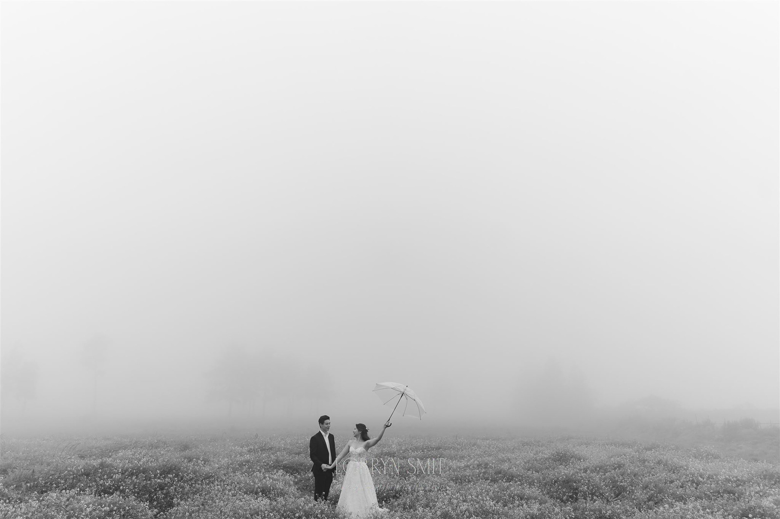 A bride holding an umbrella stands next to a groom in a flower field surrounded by fog; trees are faintly visible in the background.