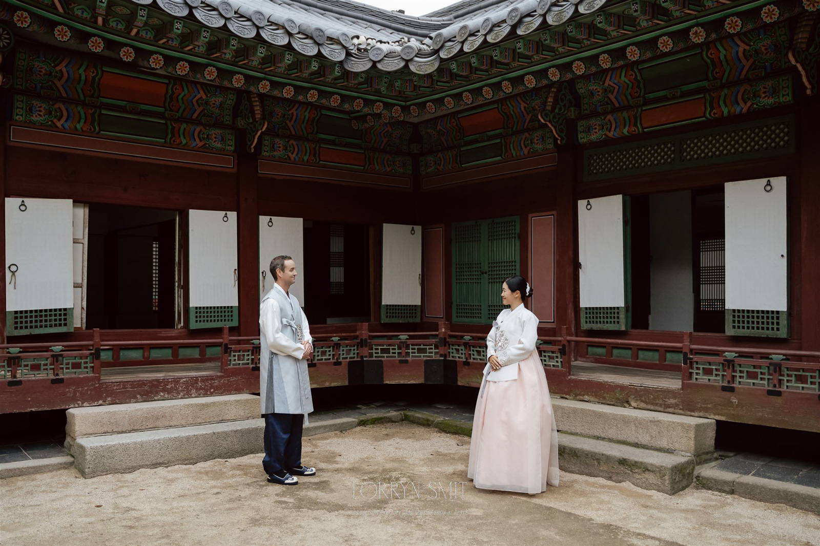 Two people in traditional Korean clothing stand facing each other in the courtyard of a historic Korean building with wooden architecture during their Korea pre wedding.