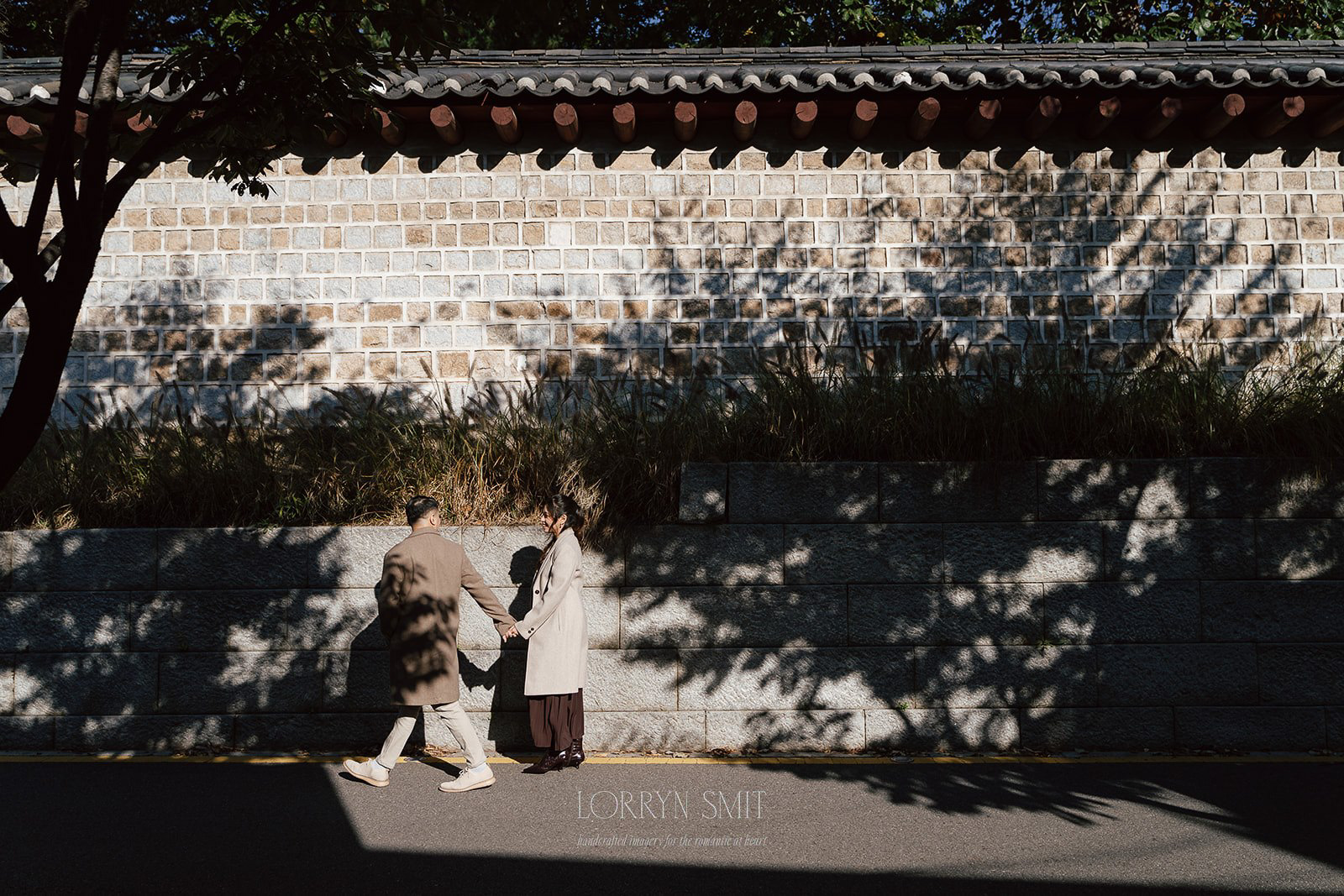 Two people holding hands walk along a stone wall in sunlight, casting long shadows on the pavement and wall behind them during their Korea pre wedding.