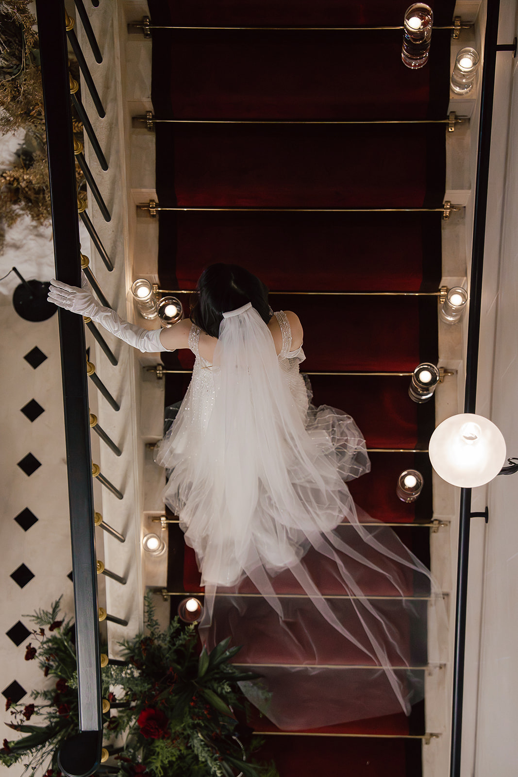 A bride in a white gown and veil descends a red-carpeted staircase, surrounded by candles and greenery, viewed from above.