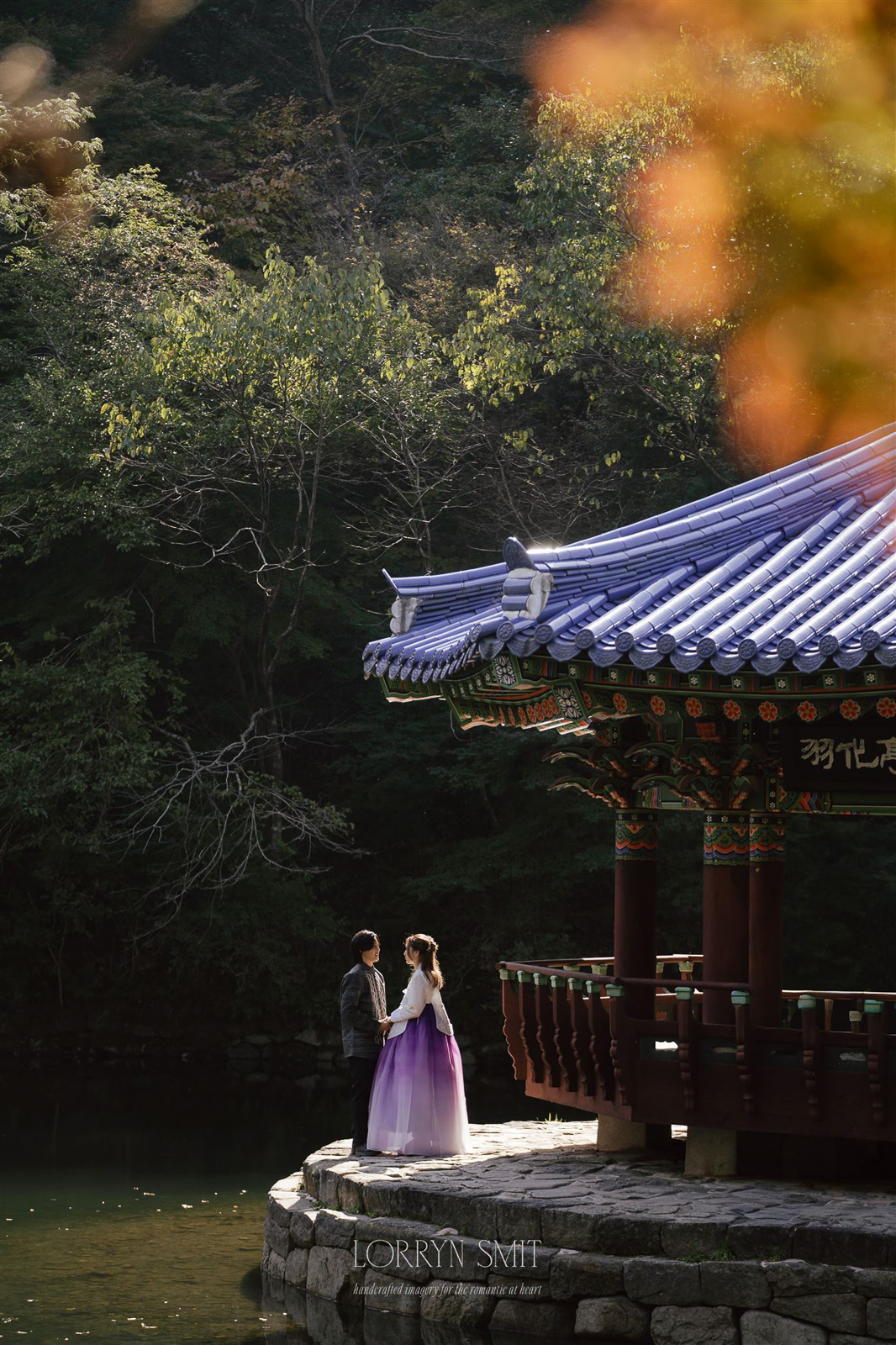 A couple stands by a traditional pavilion with a blue-tiled roof next to a pond, surrounded by trees in daylight.