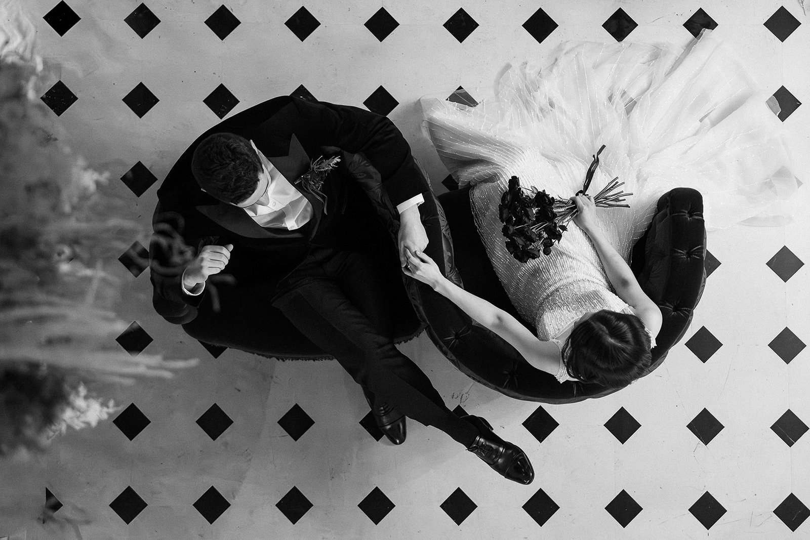 Black and white overhead photo of a bride and groom sitting on chairs, holding hands on a tiled floor with a checkered diamond pattern during their Korea pre wedding.