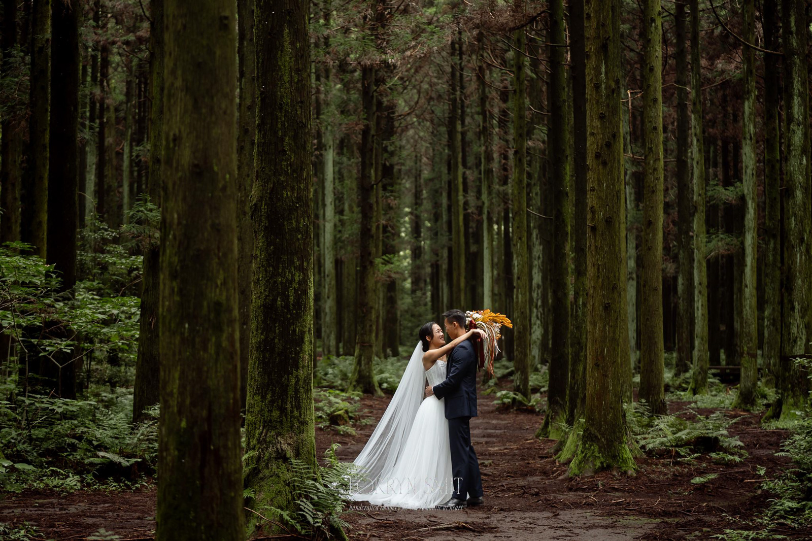 A bride and groom embrace and kiss in a forest, surrounded by tall trees and greenery during their Korea pre wedding.