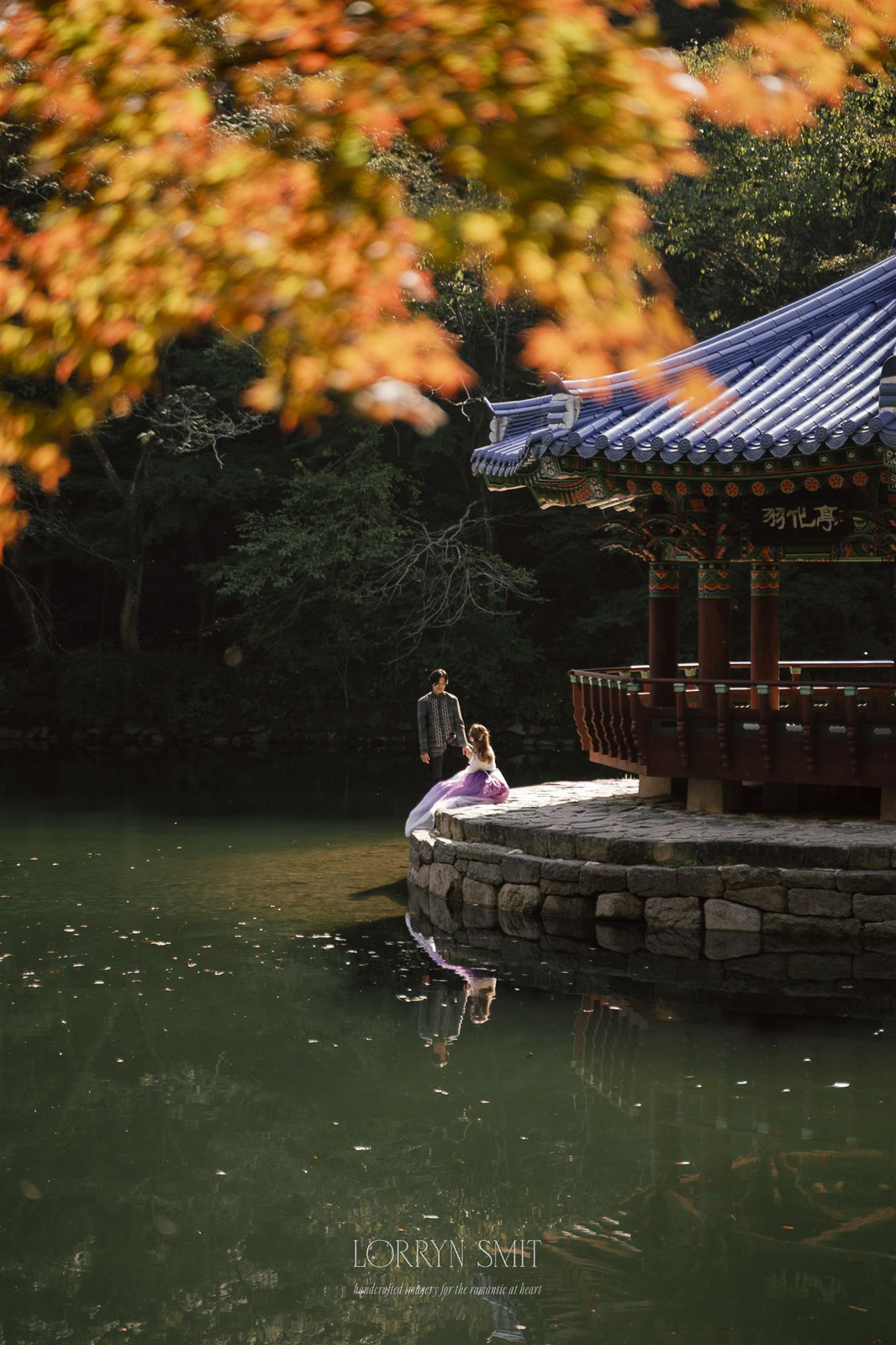 Two people in traditional Korean clothing sit by the edge of a pond near a pavilion with a blue tiled roof, surrounded by trees and autumn leaves during their Korea pre wedding.
